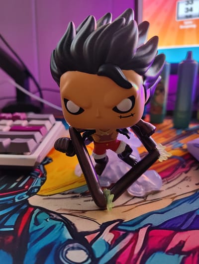 The image presents a Funko Pop-style vinyl figure of Monkey D. Luffy in his Gear 5 form, centrally positioned on a desk mat. The figure is characterized by spiky black hair, tan skin, black eyes with white pupils, and a small stitched mouth on the right side of its face. It is depicted wearing red shorts with white accents and a partially visible ripped white garment. The figure's elongated limbs are dark brown/black, consistent with the character's transformed state; one extended limb holds a thin, dark brown, curved stick-like object, while another displays a small, translucent light-green lightning bolt effect. The figure is stabilized by a clear, wavy base, simulating a cloud or smoke effect.
The desk mat beneath the figure features vibrant, stylized comic or anime artwork, primarily displaying hues of blue, red, pink, yellow, and black outlines, with abstract and partial character motifs visible.
The background, from left to right, shows a dark, indistinct object against a plain purple wall. To its right, a segment of a white mechanical keyboard with purple and grey keycaps is visible. Behind the figure's head, a computer monitor displays a digital interface with the numbers "33" and "34" in a white box, accompanied by the text "Total" and "Remaining," set against an orange-to-yellow gradient background. To the far right, two dark, cylindrical objects, likely bottles, stand against a purple wall; one object appears teal/green, the other darker. The scene is illuminated by prominent purple ambient lighting, augmented by the orange/yellow light source from the monitor.
There is no discernible information within the image to specify a geographical location, including the city of Oosterhout, Netherlands.