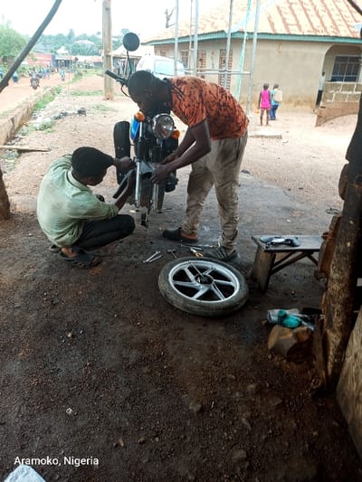 The image shows two men working on a motorcycle in a dirt lot. The man on the right is standing and using tools to remove the front wheel. The man on the left is kneeling and appears to be holding a wrench. There are other tools, a removed wheel and a small wooden stool in the foreground. In the background, there are a number of buildings, including a small house and a larger structure that appears to be a shop. The buildings have painted brick exteriors. The scene takes place in the town of Ilobu in the state of Osun, Nigeria, at the coordinates (7.835, 4.479). The ground is covered in loose gravel. There are a few trees in the distance. There are two young girls in the background, on the other side of the street. They are walking away from the camera. The day is sunny and the sky is clear. The men are dressed in casual clothing. The image was taken at a low angle.