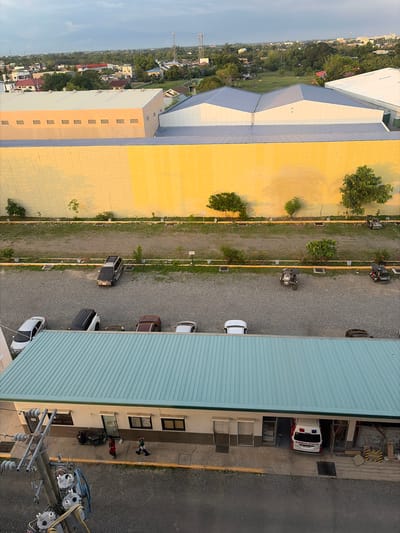 The image captures a view of an industrial area or a large complex in Dagupan City, Philippines, likely during the late afternoon or early evening due to the warm lighting and sky. In the foreground, a light blue metal roof covers a long, single-story building with a light beige facade. The building has multiple windows and doors, and an ambulance is parked inside an open bay on the right side. A utility pole with electrical equipment stands to the left of the building.
Behind this building, a wide expanse of gravel parking lot contains several vehicles, including cars, a motorcycle with a sidecar, and a scooter. A dark-colored SUV is parked further back in the lot. A row of trees and landscaping separates the parking area from a large, dominant structure.
This dominant structure is a long, warehouse-like building with a pale yellow, textured wall. Above this yellow wall, a building with a lighter colored facade and a gray metal roof is visible, featuring a distinctive V-shaped roofline. Beyond these structures, the cityscape of Dagupan unfolds, with various houses, smaller buildings, and greenery stretching towards a hazy horizon. Communication towers are also visible in the distance. The overall scene depicts a functional, possibly commercial or industrial, area within the urban landscape.