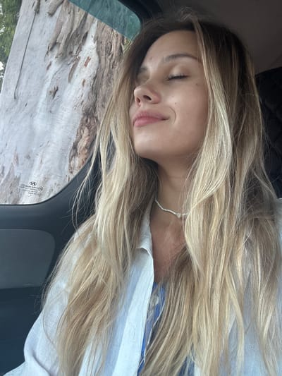 The image shows a close-up portrait of a young woman with her eyes closed and a slight smile. She has long, blonde, wavy hair that falls over her shoulders and chest. She is wearing a white button-up shirt, and a thin beaded necklace with a small heart pendant. The background shows the textured bark of a large tree, partially obscured by what appears to be a car window, with some green foliage visible. A portion of a car's interior, including a dark gray seat and a car door panel, is visible on the right side of the frame. A Hyundai logo is visible on the car window. The image is a selfie taken from a slightly low angle, suggesting the woman is in the passenger seat of a vehicle. The setting is outdoors, near a large tree, possibly during the daytime given the natural light. The location is specified as Kyndygh chyts, Abkhazia.