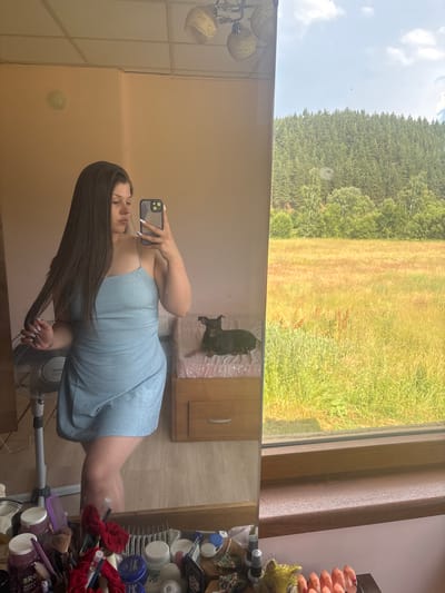 The media file shows a young woman taking a mirror selfie in what appears to be a room with a large window. The woman is wearing a light blue, sparkly dress and has long, dark hair. She is holding a smartphone in her left hand, and her right hand is touching her hair. Her expression is neutral.
The setting includes a mirror that reflects the woman and part of the room. In the reflection, a small black dog is visible lying on a pink blanket on a piece of furniture. The room also contains a fan, and to the right of the reflection, there is a shelf with various cosmetic products, brushes, and makeup items.
Beyond the room, through the window, a lush green landscape is visible, with a dense forest in the background and a field with tall grass in the foreground. The sky is bright with scattered clouds, suggesting it is daytime. The overall mood is casual and relaxed.
There are no discernible text elements in the image. The context provided, "Prodanovtsi, Bulgaria," suggests a rural or semi-rural location.