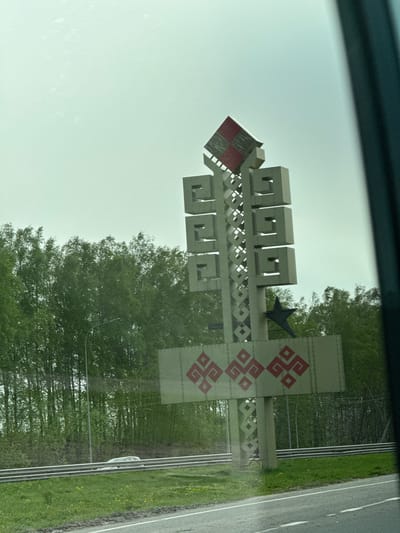 The image shows a view from inside a vehicle, likely a car. Visible outside the vehicle's window is a large monument or sign, presumably marking an entrance to a location. The structure is comprised of geometric shapes, including cube-like elements and a large, flat panel adorned with a red ornamental design. A black star is integrated into the design. The monument is set against a backdrop of green trees, suggesting it is located near a forested area or park. The sky is overcast, and a portion of a road is visible to the right of the sign. No people are visible in the scene. Based on the location, the monument is in Khyrkasy, Russia.