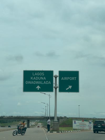A wide-angle, eye-level outdoor shot captures a highway intersection in Immigration Quarters, Nigeria. The sky is overcast and a pale blue. A large green road sign stands prominently in the center of the frame, supported by a white pole. The sign is divided into two sections. The left section, in white text on a green background, reads "LAGOS KADUNA GWAGWALADA" and has an upward-pointing arrow. The right section, also in white text on a green background, reads "AIRPORT" and has a rightward-pointing arrow.
In the foreground, on the left side of the road, a man is riding a motorcycle. He is wearing an orange t-shirt and khaki pants and appears to be looking forward. To the right of the motorcycle, a man in a white shirt and dark pants stands on the side of the road, looking towards the left.
Further down the road, a multi-lane highway stretches into the distance, with several cars and trucks visible. An overpass spans the highway in the background. On the right side of the highway, a large banner is displayed, advertising the "NIGERIAN ARMY DAY CELEBRATION 2020 GRAND FINALE". A black SUV is parked to the right of the banner. The environment appears to be a developed urban or suburban area, with paved roads, streetlights, and grassy embankments.