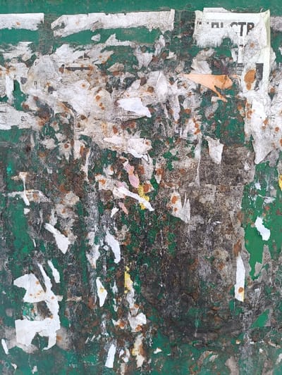 The image displays a heavily deteriorated vertical surface, identified as a public bulletin board or wall in Furmanov, Russia. The base layer is a faded, peeling dark green paint, significantly obscured by numerous irregular fragments of torn paper. These paper remnants are predominantly white and gray, with visible brown discoloration consistent with rust stains or dried adhesive. Smaller, faded sections of yellow and pink paper are interspersed. Black Cyrillic text, including characters forming "СТР" (STR), is discernible on some white paper fragments, notably in the upper right quadrant. The overall texture is rough and layered, indicative of prolonged exposure to weather and repeated poster application and removal. No individuals, direct actions, or specific interactions are depicted.