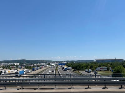 The image depicts a wide-angle view of a highway interchange in Sinsheim, Germany, on a clear, sunny day. In the foreground, a metal railing of an overpass runs horizontally across the bottom of the frame. Beyond the railing, a multi-lane highway is visible with a significant amount of truck and car traffic. On the left side of the highway, there are several commercial buildings, including trucks with "TALAY LOGISTICS" and "KFC" branding. Further into the distance, on the right side of the highway, a large stadium with a distinctive white, wavy roof structure is prominently featured, identified by signage as the Rhein-Neckar-Arena. The background consists of rolling green hills and trees under a bright blue sky with scattered white clouds. The scene appears to be a busy transportation hub with commercial and recreational facilities.