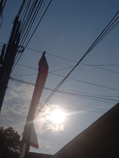 This image, captured in Bojongherang, Indonesia, presents an upward-angled view of a bright outdoor scene. The sky, predominantly clear and blue, occupies the majority of the frame, with sparse white cumulus clouds visible in the lower central area. The sun is a prominent, intensely bright point in the lower-center-right, producing a strong lens flare that radiates across the sky.
On the left side, a dark, vertical utility pole is partially visible, from which numerous black electrical and communication wires stretch horizontally and diagonally across the upper and central portions of the sky. Near the center-left, a red and white Indonesian flag hangs vertically, partially obscured by the wires. Its red stripe is positioned above the white stripe. The upper edge of a dark, angular building or structure forms a diagonal silhouette along the bottom right of the image. In the bottom-left corner, a dark mass of tree foliage is visible in silhouette. No people are present. The scene is illuminated by direct sunlight.