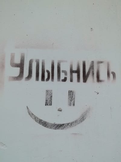 The image shows a stencil of a smiley face and the Russian word "УЛЫБНИСЬ" (which translates to "Smile") spray-painted on a pale blue, textured wall. The stencil is black and appears to be applied with spray paint, with some overspray visible around the edges. The smiley face has two square eyes and a curved mouth. The letters are blocky and somewhat uneven due to the stencil application. The background is a plain, lightly textured wall. There are no people or other objects visible in the immediate vicinity. The scene is devoid of any specific Pinsk landmarks.
