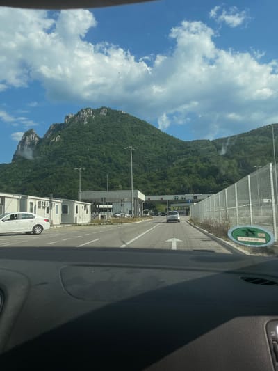 The image depicts a view from inside a vehicle looking out towards a road and a mountainous landscape. The road appears to be a toll plaza or a border crossing, with several lanes indicated by white arrows and road markings. There are some vehicles visible on the road, including a white van in the foreground on the right.

Adjacent to the road are several modular buildings, possibly administrative offices or waiting areas. Beyond these buildings, a dense, green mountainside rises steeply, covered in trees. The sky is blue with fluffy white clouds, suggesting a clear day.

A circular green sticker with some text and a symbol is affixed to a fence or barrier on the right side of the image. The text on the sticker is partially obscured but appears to be in a non-Latin script. There are also signs with arrows and symbols above the lanes on the road, indicating directions or lane usage.

The overall scene suggests a journey through a scenic, mountainous region, possibly during daylight hours given the brightness and clear skies. The presence of the toll plaza or border crossing infrastructure indicates a point of transit.