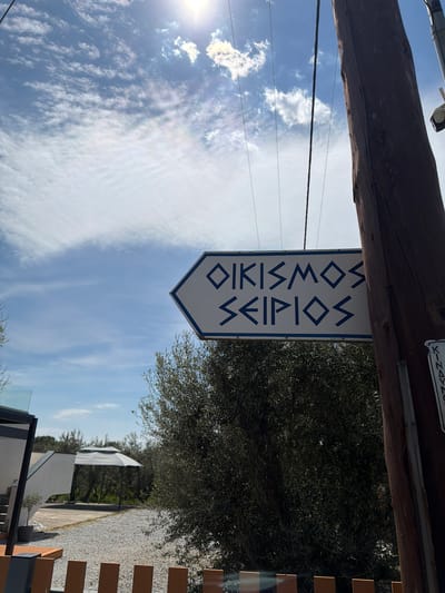 A low-angle, daylight shot shows a sign with the Greek words "OIKISMOS SEIKIOS" (Οικισμος Σεικιου) in dark blue lettering on a white background with a dark blue border, which is hanging from a weathered wooden beam. The sign points downwards, suggesting a direction. Behind the sign, a partly cloudy blue sky fills much of the frame. To the right of the sign, dense green foliage is visible. In the lower right corner, a wooden fence and glimpses of buildings and a graveled path appear. The photograph is taken outdoors and the lighting suggests a sunny day. The location is likely near the Seikios settlement in Megas Alexandros, Greece, as indicated by the sign's inscription. There are no people visible.