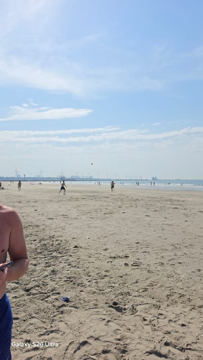 This is a vertically oriented, full-shot photograph of a beach scene at Hook of Holland, Netherlands. The image is dominated by a wide expanse of sandy beach in the foreground and midground, with faint footprints visible in the sand. In the distance, the sea meets the shore, and further beyond, numerous wind turbines dot the horizon, suggesting a windy coastal environment. The sky is a clear, pale blue with wispy white clouds.

Several people are visible on the beach, scattered across the sand and near the water's edge. Some appear to be individuals walking, while others seem to be playing or engaged in activities. One person is particularly prominent in the midground, running with their arms outstretched. There's also a small dark object in the air, possibly a ball, suggesting a game is in progress. A person's arm and shoulder are visible in the bottom right corner of the frame, holding a phone.

The lighting suggests it is daytime, likely with bright sunshine given the clear sky and the clear shadows cast on the sand. The overall mood appears relaxed and leisurely, consistent with a day at the beach. The presence of wind turbines is a strong indicator of the location being a coastal area in the Netherlands.

Visible text in the image, located in the bottom right corner, reads "Galaxy S26 Ultra".