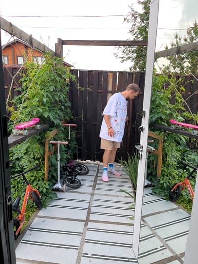 A man stands in an outdoor area in Sukhaia Balka, Russia, looking down. He is wearing a white t-shirt with a floral pattern, tan shorts, pink slip-on shoes, and white socks with blue stripes. The area appears to be a patio or walkway made of grey tiles with black and white stripes. To his left, a wooden fence and lush green vegetation are visible. Resting on a wooden beam above the vegetation are two pink skateboards. Several scooters are parked on the ground, including an orange one and two smaller silver and black ones. A large mirror is positioned to the right of the man, reflecting the scene. In the background, a wooden fence borders the area, and a two-story wooden house with windows is visible beyond it, partially obscured by trees. The sky is overcast.