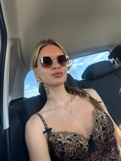 An adult female, characterized by a light skin tone, long blonde hair styled in a single side braid, is depicted seated in the rear passenger compartment of a vehicle. She wears hexagonal-framed sunglasses with dark lenses, large ornate gold-toned earrings, and a thin silver-toned chain necklace featuring a key-shaped pendant. Her attire consists of a black and gold patterned top or camisole with lace trim and small black bow accents on the straps. A black snake-design tattoo is visible on the left side of her neck. Her lips appear glossy. The vehicle's interior features black leather or synthetic seating and a light-colored headliner. Through the rear window, a blue sky with scattered clouds is visible, indicating daylight conditions. The woman is positioned with her torso angled slightly to the right, facing directly towards the camera. The scene is depicted as occurring in Granadilla de Abona, Spain.