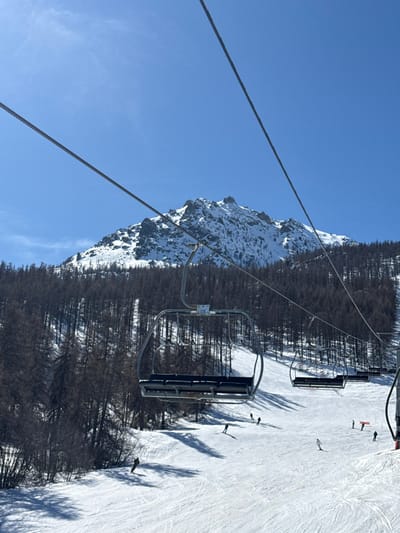 The image presents a winter ski resort scene in Saint-Chaffrey, France, under a clear, bright blue sky. Two prominent parallel steel cables, part of a ski lift system, extend diagonally across the upper frame from left to right. Several empty ski lift chairs are visible, suspended from these cables, with one chair, labeled "126," centrally positioned, featuring four black padded seats. In the background, a snow-covered mountain peak with exposed rock formations rises against the sky. Below the mountain and extending across the mid-ground is a dense forest composed of dark, leafless trees, partially covered in snow. The lower half of the image displays a wide, groomed, snow-covered ski slope, exhibiting numerous ski tracks and distinct, elongated shadows cast by skiers and unseen objects. Approximately ten individuals, identified as skiers, are observed descending the slope, scattered across its surface. Two small orange safety markers or poles are discernible on the right side of the ski slope.