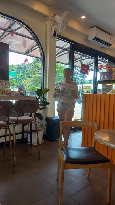 A horizontally oriented photograph captures a scene inside what appears to be a cafe or restaurant in Plai Phraya, Thailand. The cafe's interior is well-lit, with natural light streaming through large windows that offer a lush, green outdoor view.

A woman stands near the window, her image visible through the glass. She wears a striped top and denim shorts. She is holding two coffee cups, one in each hand, and appears to be posing for the picture.

The foreground features cafe furniture, including a set of chairs with brown upholstery and metal frames, and a table with a round wooden top. A decorative plant in a white pot stands near the chairs. In the background, a closed sign can be seen, along with a menu display. Overhead, a white wall-mounted air conditioning unit and fan are visible. The overall impression is of a relaxed and casual cafe setting.