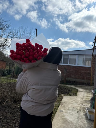 An individual, seen from behind, holds a large bouquet of numerous red tulips wrapped in white translucent material. The person wears a light beige puffer jacket and a dark head covering. The scene is outdoors under a blue sky with scattered white clouds, located in Trudobelikovskiy, Russia. In the background to the left, bare tree branches and a partial brick structure are visible. To the right, a single-story building with a red brick facade and white-framed windows stands. A concrete path runs through the foreground, bordered by bare soil and sparse dry vegetation. A dark pole, a white rectangular basin containing liquid, and a green hose are visible on the far right.