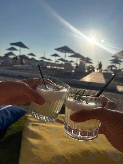 Two hands are holding up glasses of drinks with frothy tops and rosemary sprigs, clinking them in a toast at a beachside location in Foça, Turkey. The sun is shining brightly in the background, with a lens flare visible. Beach umbrellas and lounge chairs are set up on the sand, and a wooden walkway extends towards the sea. The overall scene evokes a relaxed and celebratory atmosphere, likely at sunset given the warm lighting.