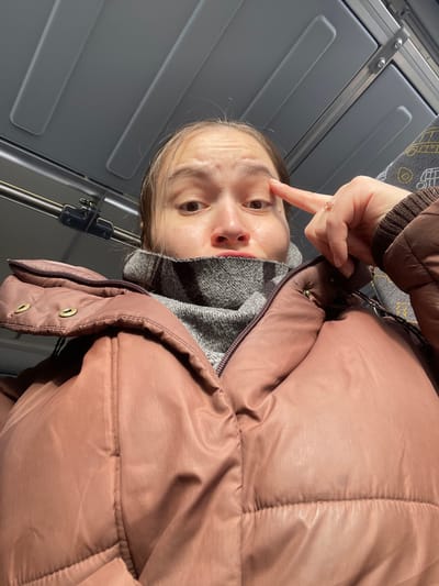 The scene depicts the interior of a public transport vehicle in Fryazevo, Russia. A female individual is centered in a low-angle, close-up shot, looking directly at the camera with wide-open eyes and slightly parted lips. She is dressed in a dusty rose-colored puffer jacket over a grey, textured high-neck garment. Her left hand is raised, with the index finger pointing towards her left eye; a ring is visible on her left ring finger. The background consists of a light grey paneled ceiling, a horizontal metal grab rail, and a segment of patterned upholstery, likely a seat.