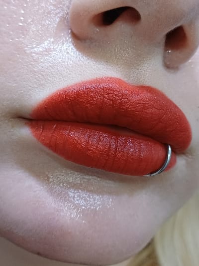 The scene depicts a close-up, static portrait of a person's lower face. The subject's lips are coated entirely with a matte, vibrant red lipstick. A silver-toned, circular metal piercing is inserted through the left side of the lower lip (viewer's right). The surrounding skin, visible on the philtrum, above the upper lip, and below the lower lip, exhibits a fair complexion with discernible pores and a subtle sheen, possibly from a cosmetic product or natural skin oils. The lower part of the nose, including the nostrils, is visible at the top of the frame. A small section of light-colored hair is present at the bottom right periphery. The image is tightly cropped, providing no discernible background or environmental context. The contextual location is in the city of 서대문구, South Korea.