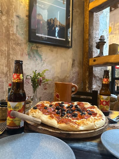 A close-up, medium shot captures a table setting in what appears to be a rustic restaurant in Zacatecas City, Mexico. The foreground is dominated by a freshly baked pizza with pepperoni and black olives, placed on a wooden cutting board. To the left of the pizza stands a brown glass bottle of Dos Equis beer, its label clearly visible. Another bottle of Dos Equis is positioned to the right, slightly further back. Between the pizza and the first beer bottle, a small vase holds a sprig of greenery with a few dried pink flowers. A rustic, orange-brown ceramic mug with a handle rests behind the pizza. The table surface is made of weathered, multi-colored wood. In the background, the wall features a textured, distressed plaster with hints of green and brown paint. A framed picture of a mountainous landscape hangs on the wall. To the right, a wooden beam supports a shelf displaying a dark, antique-looking object, possibly a lamp or decorative piece, and another cylindrical container. The overall ambiance suggests a casual dining experience, characteristic of many eateries found within the historic center of Zacatecas City.