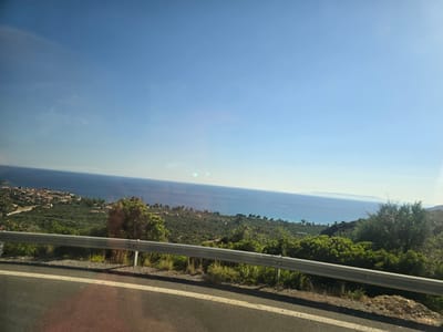An elevated view captures a coastal landscape under a clear, bright blue sky. In the foreground, a paved road with white markings curves to the left, bordered by a silver metal guardrail. Beyond the guardrail, a green, undulating terrain is dotted with trees and vegetation. In the mid-ground, a village with terracotta-roofed buildings is nestled along the coastline. The sea stretches out in the background, meeting the horizon where a distant landmass is faintly visible. The scene is likely a scenic overlook on a sunny day in the Municipal Unit of Lefktro, Greece, offering a panoramic view of the sea and the coastline.