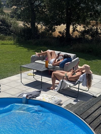 The image depicts an outdoor scene with two individuals relaxing in a backyard setting. In the foreground, a blue swimming pool covered with a solar blanket is visible. To the right of the pool is a wooden deck. Behind the pool, a tiled patio area is laid out.
On the patio, a dark, rectangular coffee table sits between two seating arrangements. On the table, a bright orange water bottle is placed. Yellow flip-flops are visible underneath the table.
Further back on the patio, one person is reclining on a grey outdoor sofa, holding a smartphone and looking at it. They are wearing a white top and light-colored shorts. Another person is seated in a white lounge chair, also looking at a smartphone. This individual is wearing a green bikini and a checkered hat. A towel is draped over the back of the lounge chair.
The background shows a well-maintained green lawn, mature trees with dense foliage, and beyond that, a dry, agricultural landscape under a clear blue sky. The lighting suggests it is a sunny day. There is no indication of the location being in San Lorenzo de Tormes, Spain, or any other specific geographical location.