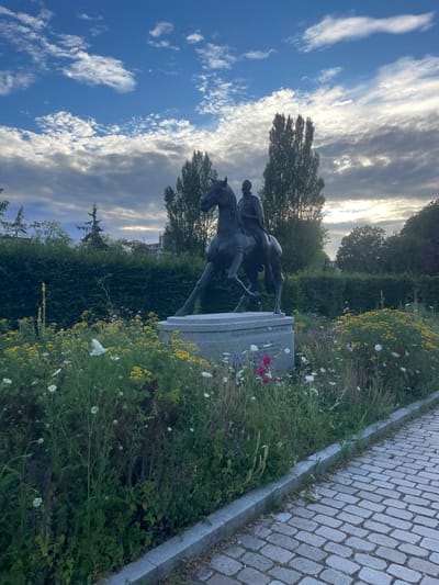 A statue of a man on horseback is depicted in an outdoor setting, possibly a park or a public square. The statue is dark in color and appears to be made of bronze. The horse is depicted in mid-stride, with the rider seated upright. The base of the statue is a grey stone monument, on which some text is visible.
The scene is surrounded by lush greenery, with various plants and flowers growing around the base of the monument. Some of the flowers are yellow and white, adding a touch of color to the scene. In the background, there are trees and a hint of buildings, suggesting an urban or semi-urban environment. To the right of the statue, a paved pathway made of cobblestones is visible.
The sky above is a mix of blue and white, with dramatic clouds that suggest either dawn or dusk. The sun appears to be low on the horizon, casting a soft light on the scene. The overall mood is serene and historical, with the statue serving as a focal point of remembrance.
The visible text on the monument reads "Prins Van Montmorency Graaf van Horne". This text indicates that the statue likely commemorates Prince van Montmorency, Count of Horne, a historical figure. The location context provided is Weert, Netherlands, which is consistent with a monument to a Dutch nobleman.