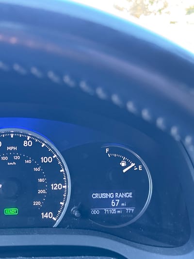 The image displays the instrument cluster of a vehicle, observed from the driver's perspective, located in Rancho Palos Verdes, United States. A dark-colored steering wheel with visible stitching partially occupies the upper portion of the frame. The illuminated instrument panel features a speedometer on the left, calibrated from 0 to 140 MPH (and 0 to 220 KM/H), with a green "READY" indicator light active at the 7 o'clock position. On the right side, a fuel gauge indicates that the fuel tank is approximately one-quarter full. Below the fuel gauge, a digital display presents a "CRUISING RANGE" of "67 mi," an odometer reading of "71105 mi," and an "OUTSIDE" temperature of "77°F." The background above the dashboard is brightly lit and blurred, suggesting an outdoor, daylight environment. No individuals are visible.