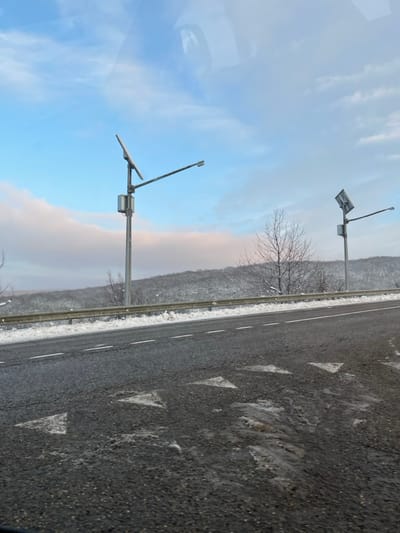 The image presents a forensic view of a winter roadside scene in Gostagaevskaya, Russia. The foreground consists of an asphalt road with a broken white lane line and multiple white triangular markings pointing towards the viewer. The road surface displays patches of moisture and melted snow/ice. A silver-colored metal guardrail spans the length of the visible road, positioned to the right of the lane line, with snow accumulated against its base. Two utility poles are visible; the left pole, prominent and closer, features a solar panel angled upwards, a grey cylindrical battery or control box, and a horizontal arm extending left, terminating in a streetlight fixture. A second, similar utility pole is visible further back on the right, also equipped with a solar panel and streetlight. Behind the guardrail, the terrain slopes upwards, covered in snow-dusted deciduous trees with bare branches. The sky is clear blue with scattered white and faint pinkish clouds. No individuals are present. Faint, indistinct reflections, consistent with a vehicle's interior, are visible in the upper portions of the image, suggesting the photograph was captured from within a vehicle.
