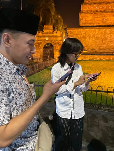 Two individuals are visible at night in Kauman, Indonesia, specifically in front of the illuminated Great Mosque of Kauman. The subject on the left, partially framed, is a male wearing a black songkok and a patterned short-sleeved shirt, holding a smartphone in his right hand and looking down at it with a slight smile. A light-colored sling bag is visible across his body. The subject on the right is a male with long dark hair and black-rimmed eyeglasses, dressed in a white long-sleeved button-up shirt with black trim and dark patterned pants. He is holding two smartphones, one in each hand, and intently focused on the screen of the phone in his right hand. A black sling bag strap is visible over his left shoulder. Both individuals are looking down at their respective phones.

In the background, a large brick structure, characteristic of the Great Mosque's walls and an arched gateway, is brightly illuminated with warm orange light. A section of manicured green grass, also orange-lit, is visible behind a dark metal fence that separates it from the paved foreground where the individuals stand. Overhead, dimly lit tree foliage is discernible against the dark night sky. The ground beneath the subjects appears paved and reflects some light.