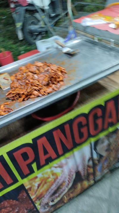 The image shows a close-up of a food stall in Sukodono, Indonesia. The foreground is dominated by a large, flat metal griddle with a pile of cooked squid, coated in a reddish-orange sauce. A spatula rests on the griddle to the left of the squid. Below the griddle, a brightly colored banner with large red letters that read "JIPANGGA" is visible. The banner also features images of seafood. In the background, a blurry motorcycle wheel and a red plastic container are present. The overall scene suggests a bustling street food vendor.