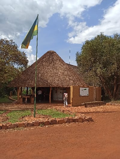 A thatched-roof building stands adjacent to a red-dirt path, bordered by a low stone wall and green vegetation. A flag, predominantly green and yellow, flies to the left. A person is standing in front of the building, near a sign. A large tree with dense foliage grows next to the building. The sky above is blue with scattered clouds.