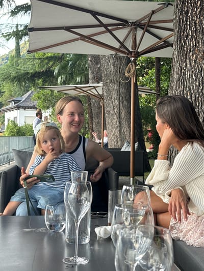 In Pognana Lario, Italy, three people are seated at an outdoor table beneath a large umbrella. A blonde woman on the left holds a young child on her lap, who is looking at a smartphone. The woman is smiling and looking to her right. Seated to her right is another woman wearing a cream-colored sweater and a pink skirt. She is looking away from the camera and resting her chin on her hand. A partially visible person is seated in the background to the right. The table in front of them is set with multiple wine glasses, some containing white wine, and a carafe. The background shows lush green trees and a building.