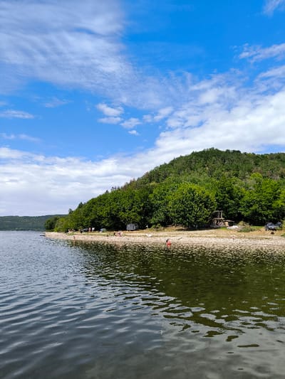This image depicts a Dniester River or Reservoir shoreline scene near the city of Horaivka, Ukraine, under a bright blue sky with scattered white cumulus clouds. The foreground is dominated by rippled, murky green-grey water. A light-colored, pebbly beach extends across the midground, bordering a densely forested hillside that rises steeply from the water's edge and extends into the background.
Approximately 10-15 individuals are dispersed along the shoreline and in the water. On the beach, multiple tents are pitched, including at least two grey and one lighter-colored tent. A rudimentary wooden structure is present on the cleared ground further right from the tents, and a dark-colored SUV is parked beneath the trees at the far right. Several people are observed standing or sitting on the beach. At least two individuals are partially submerged in the water near the shore. One person is seated in a red kayak located in the water on the right side of the frame.