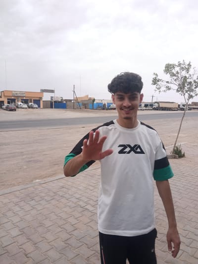 A young man with dark, curly hair and a mustache is standing on a paved walkway in an outdoor setting. He is wearing a white t-shirt with black and green accents and black pants. He is raising his right hand, palm facing forward, in a waving gesture. The background shows a road with several cars parked and some large trucks in the distance. Behind the road, there are buildings, including a light orange building with Arabic text above the entrance, and a blue building. There is a thin tree with green foliage to the right of the man. The sky is overcast. The location is identified as the city of Hun, Libya.
