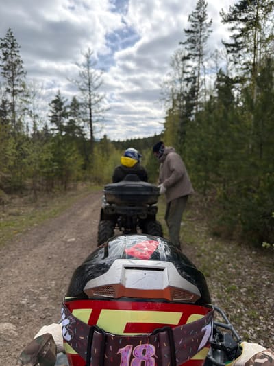 The scene is an outdoor woodland environment. A person wearing a helmet with red and yellow stripes is in the foreground, likely on an ATV or four-wheeler. In the background, two individuals are visible, one wearing a coat and another wearing a blue helmet with a yellow covering. The ATV is on a dirt or gravel path, and trees line the path. The sky is cloudy. The city is not mentioned.