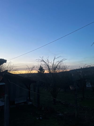 The image depicts an outdoor scene at twilight in Topchii, Bulgaria. The sky gradient transitions from a deep blue overhead to a lighter blue, culminating in a yellow-orange band at the horizon, consistent with either sunrise or sunset. A thin, dark cable or wire extends diagonally across the upper portion of the frame from the upper left to the mid-right. In the foreground, a section of a tiled roof is visible on the far left, adjacent to a dark structure featuring a corrugated metal roof. A chain-link fence, supported by dark posts, runs horizontally across the midground. Beyond the fence, several leafless deciduous trees are silhouetted against the brighter horizon, with dark, rolling hills visible in the distant background. A terracotta pot is observed hanging from a fence post or branch in the mid-right. On the ground near the lower left fence line, two small, light-colored containers are present. The ground is covered in dark vegetation. No individuals or explicit activities are depicted.