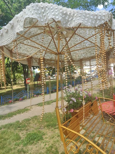 A golden decorative swing set with a large, ornate umbrella stands outdoors on a grassy area in Kryvyi Rih, Ukraine. The umbrella is made of white fabric with intricate lace and adorned with strings of gold and silver beads, as well as artificial roses. Beneath the umbrella, a planter box filled with pink and purple petunias is mounted on the swing. The swing itself is golden and features a slatted seat. In the background, a blue decorative fence with ladybug motifs separates the swing area from a path and a play area with a small wooden structure. Trees and a partly cloudy sky form the backdrop.