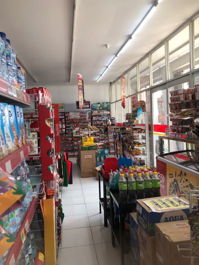 This is a medium-shot, eye-level interior photograph of a minimarket in Pilang, Indonesia. The store is well-stocked with various products displayed on shelves and racks. In the foreground, to the left, are shelves with bottled water and boxed drinks. In the center, a red display rack holds colorful shopping bags. Further back, more shelves are filled with packaged food items, beverages, and household goods. The floor is tiled, and fluorescent lighting illuminates the space. The background shows the entrance to the store, with large glass doors and windows. There are no people visible in the image. The overall impression is of a clean and organized retail environment.