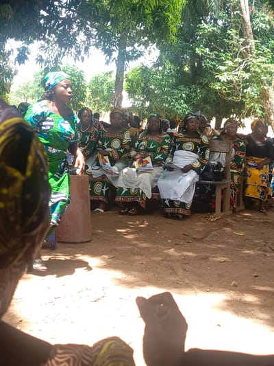 The image shows a gathering of women in a rural setting, likely in or near Ninzo South, Nigeria. Most of the women are wearing matching outfits with green, white, and brown patterns. The text "ECWA ZUMUNTA," "CAR MATAN," and "UMUNTIR ITAUNA" are visible on their dresses. Some women are seated on wooden benches, holding books or papers. One woman stands near a concrete post, addressing the group. The ground appears to be dirt or packed earth. Foliage from trees provides shade. The foreground is somewhat obscured, suggesting the photographer is part of the crowd.