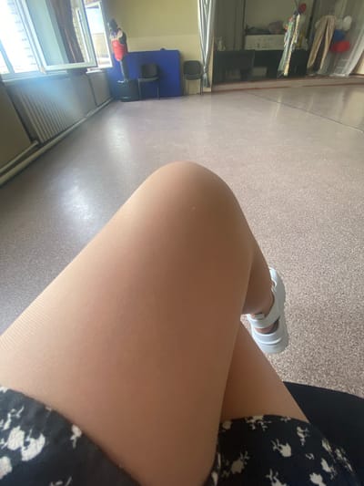 The image shows a close-up view of a person's legs and feet, sitting on what appears to be a chair. The person is wearing sheer pantyhose and white platform sandals. A portion of a dark dress with a floral pattern is visible at the bottom left. The background suggests an indoor location, possibly a school or community hall in the city of Primorsky, Russia. The floor is a light speckled linoleum. In the distance, a punching bag with a red vest is visible near a window with curtains. Other objects like chairs and a blue mat are also present in the background. The lighting appears to be natural, coming from the windows.