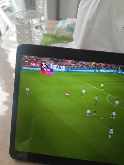 An electronic tablet displays a live football match between Manchester United (MUN) and Aston Villa (AVL), with the score MUN 3 and AVL 1. The match clock shows 98:06, with an additional 8 minutes of added time indicated. The screen depicts multiple football players on a green pitch; some wear red kits, others wear white or light blue kits, and one referee is visible in a black and yellow kit. A football is present near a cluster of players. A blurred stadium crowd occupies the background of the displayed match, above digital advertising banners that include "malaysiaairlines." The tablet rests on a light-colored, possibly wooden, surface. To the left of the tablet, a clear plastic water bottle, partially filled with water, is situated. To the right, a white tissue box or pile of tissues is visible, partially obscured by a green object. The background beyond the table is out of focus, consistent with an indoor domestic or common setting. The precise location within Cherwell District, United Kingdom, is not discernible from the visual evidence provided.