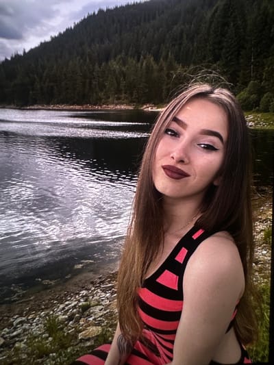 A young woman is posing for a selfie at the edge of a lake. She has long, light brown hair, dark eyebrows, and is wearing dark lipstick. She is wearing a black and pink striped top. The background shows a dark lake with choppy water and a dense, dark green forest on a hillside. The ground in the foreground is covered with pebbles and some sparse green vegetation. The sky is overcast with clouds.