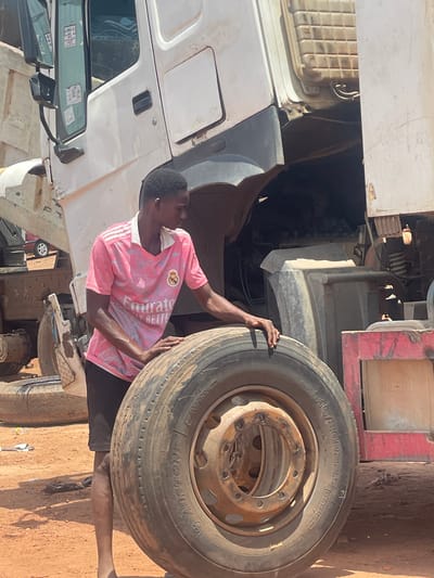 A man in a pink Real Madrid jersey stands next to a large truck tire. The truck's front is visible, and the surroundings appear to be outdoors on a dirt-covered surface. The tire's sidewall has "315/80R22.5" and "AUSTONE" imprinted. The location is Kwali, Nigeria.