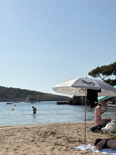 The image shows a sunny day at a beach in Sant Joan de Labritja, Spain. The perspective is from the beach, looking out towards the sea. There are several people visible. On the sand, a woman in a bikini is lying down under a white umbrella with a lizard logo and the word "Ibiza" written on it. Further down the beach, another person is partially visible, also lying down. In the water, a man is standing. The sea is calm and the sky is clear blue, suggesting it is midday with good weather. Boats are docked in the water along the coastline, which is lined with trees and rocky outcrops. The sand is light brown and appears to be typical beach sand. The overall mood is relaxed and typical of a vacation at the beach.