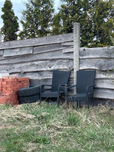 The image displays an outdoor scene in Самахвалавіцкі сельскі Савет, Belarus, featuring a weathered wooden fence with various items stored against it. The fence, constructed from rough, greyed timber planks, stands vertically. A horizontal, rectangular concrete beam is affixed to the fence at mid-height.
Against the fence, two black outdoor chairs, made of a woven material, are stacked. Below them, two black car tires with distinct treads lean against the wooden structure. To the right of the tires, a pile of reddish-brown bricks is neatly stacked on the ground.
The ground area is covered with a mix of green and dry, straw-like grass, suggesting an unkempt patch or an area in transition between seasons. In the background, dense coniferous trees with dark green foliage are visible under a light, possibly overcast, sky. The light indicates it is daytime. No people are present, and no specific activity is taking place. There is no visible text within the image. The scene portrays a utilitarian outdoor space, possibly a backyard or a storage area.