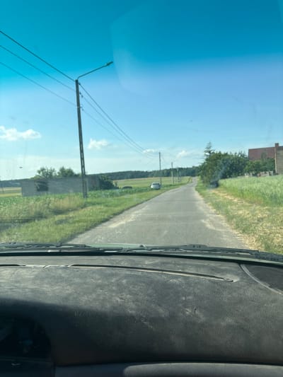 An outdoor, eye-level, point-of-view shot captures a dusty car dashboard in the foreground, looking out onto a rural road in Remiszówka, Poland, under a bright blue sky with scattered clouds. The road, paved with asphalt, winds through a landscape of green fields and sparse vegetation. A concrete utility pole with power lines stands to the left of the road, near an unfinished concrete structure. In the distance, trees form a dark line along the horizon, and a red-brick building is visible to the right. A silver car is parked on the left side of the road, further down. The sun is bright, indicating daytime.