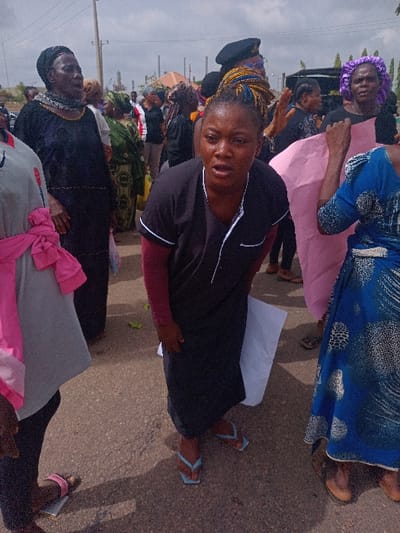 The image shows a group of people gathered on a paved street in Ungwan Tanko, Nigeria. At the center of the image is a woman wearing a black long-sleeved shirt, a black skirt, and blue flip-flops. She is standing with a distraught expression and her hands clasped in front of her. The street is littered with people, some wearing traditional clothing. In the background, there are several tall structures, potentially industrial, and a cloud-filled sky. The image appears to capture a protest or demonstration. A woman in the foreground, wearing a blue and white patterned skirt, holds a pink poster. There is a woman on the left in a long grey shirt with a pink scarf, and a woman in the background, wearing a black dress and hijab, is holding a sign. The image depicts a scene of frustration and potential confrontation in a public area.