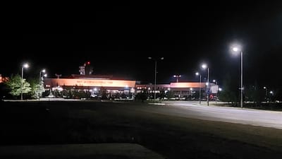 The image shows a nighttime view of Kurylys Airport, located in Kurylys, Kazakhstan. The airport building is illuminated, with large orange panels on its facade displaying the text "KURYLYS-AIRPORT." Several streetlights line the road in front of the airport, casting a glow on the asphalt. The surrounding area is dark, with trees visible on the left and right sides of the frame. A few vehicles are parked in front of the building. The sky is black, indicating it is nighttime, and there are no visible people in the image. The scene is static and appears to be a wide shot of the airport terminal and its immediate surroundings.