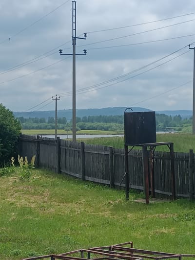 The image depicts an outdoor scene in Granovshchina, Russia, featuring a wooden fence in the foreground, separating a grassy yard from a body of water and a tree-lined landscape in the background. Power lines traverse the sky, supported by utility poles. On the right side of the image, a dark, rectangular water tank is mounted on a metal stand. The sky is overcast.