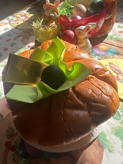 The image shows a table arrangement, likely indoors, on a tablecloth printed with Easter-themed imagery, including rabbits and flowers. The main focus is a large, braided sweet bread, possibly a holiday loaf. A folded, light green napkin and a gold-wrapped "Merci" chocolate bar are placed on top of the bread, with a dark, oval object (perhaps an Easter egg or a decoration) nestled within the napkin's folds. Adjacent to the bread, on the left side of the image, is an assortment of Easter decorations, including painted eggs, a ceramic bunny figurine, and a decorative egg with straw-like accents. The lighting is bright and sunny, casting shadows across the scene. The location in Tompsan, Bulgaria, cannot be confirmed from the image alone.